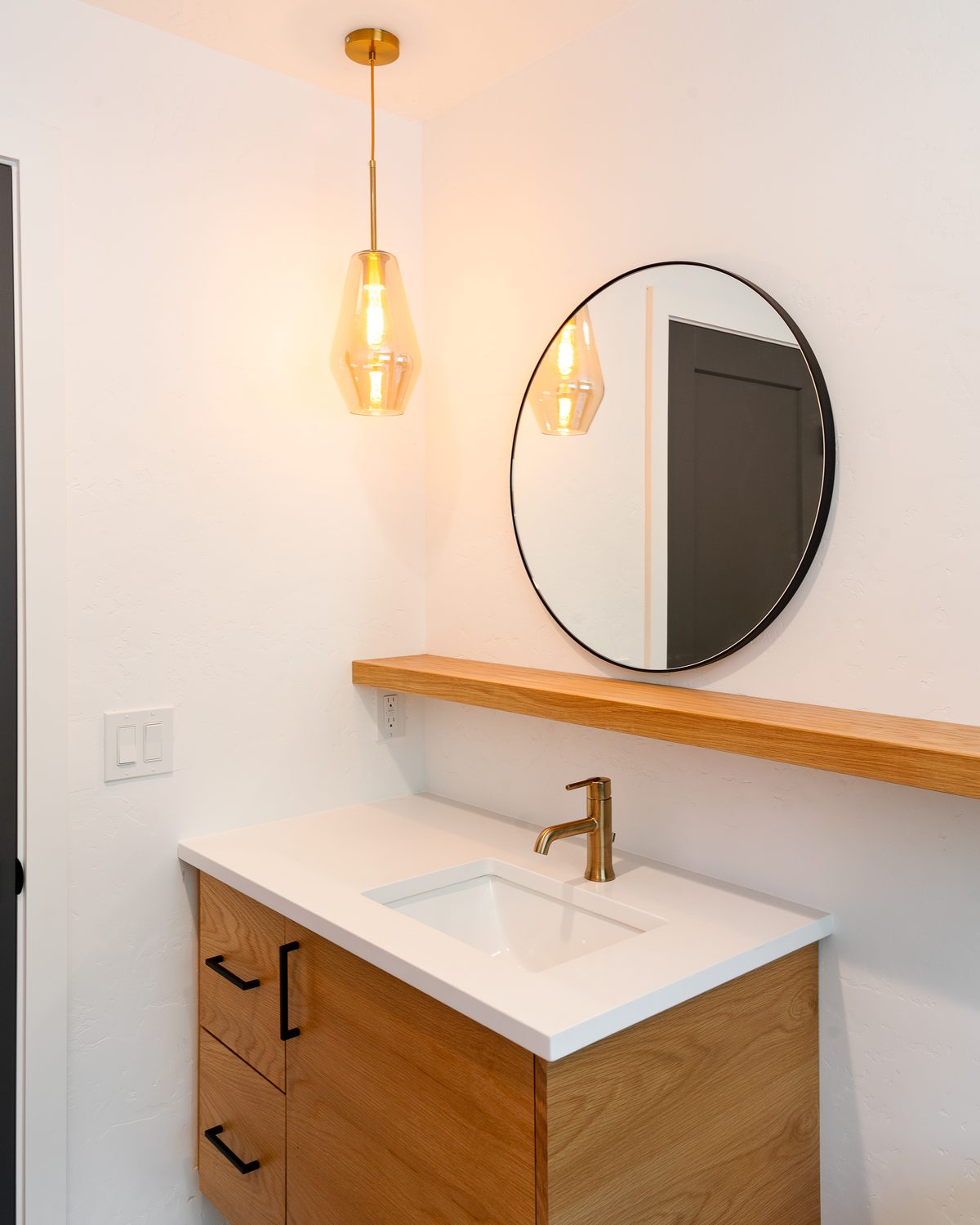 Floating white oak single vanity with a brass faucet, round black-framed mirror, oak shelf, and amber glass pendant