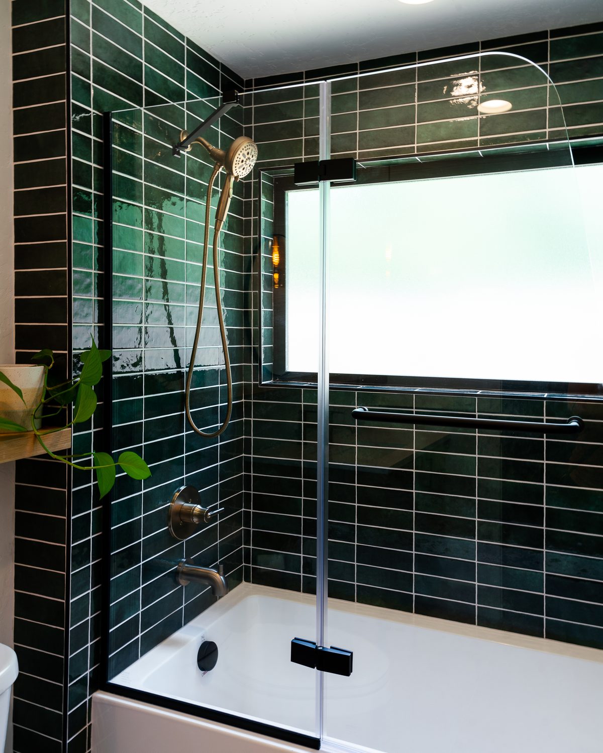 Tub and shower with glossy dark green stacked tile, a brushed handheld shower, and a black-framed hinged glass panel