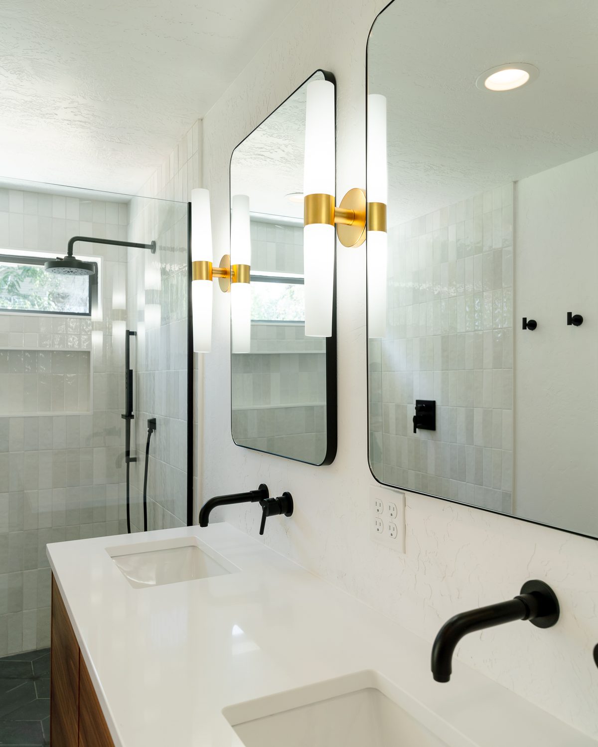 Walnut vanity with a white quartz top, black wall-mount faucets, brass sconces, and a curbless shower with a black rain head beyond