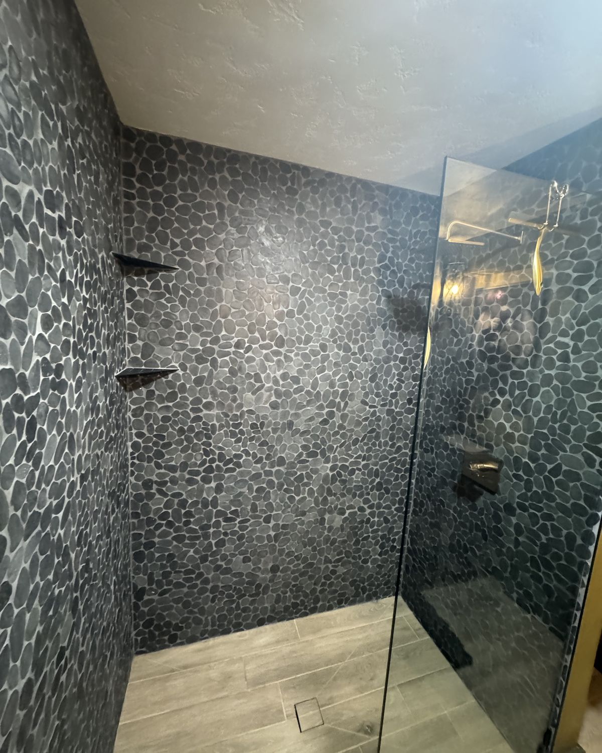Shower clad in dark pebble stone tile with stone corner shelves and a frameless glass panel