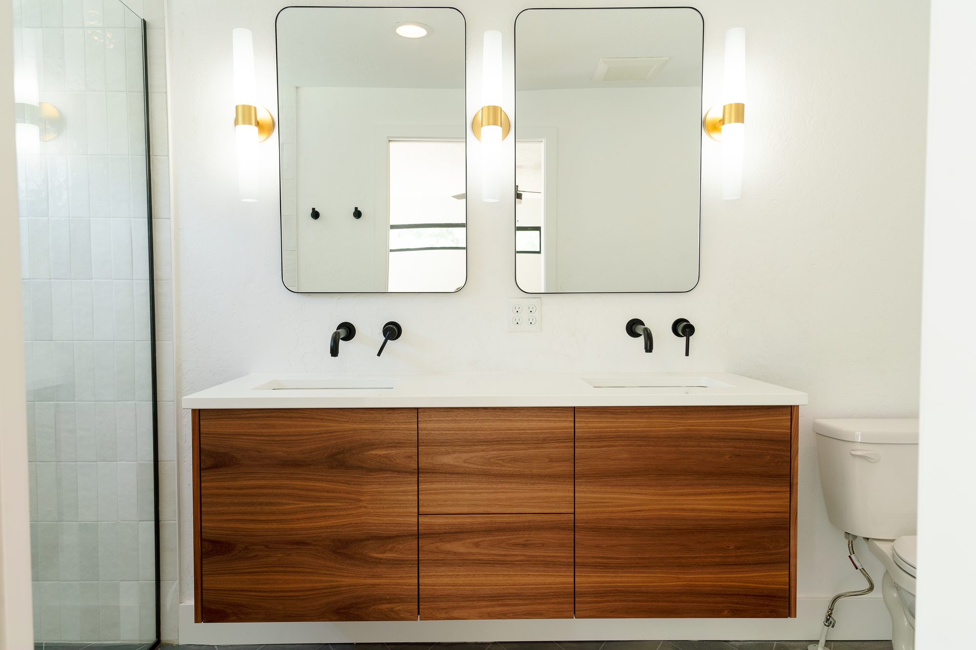 Floating grain-matched walnut double vanity with a white quartz top, black wall-mount faucets, two black-framed mirrors, and brass sconces, beside a glass shower