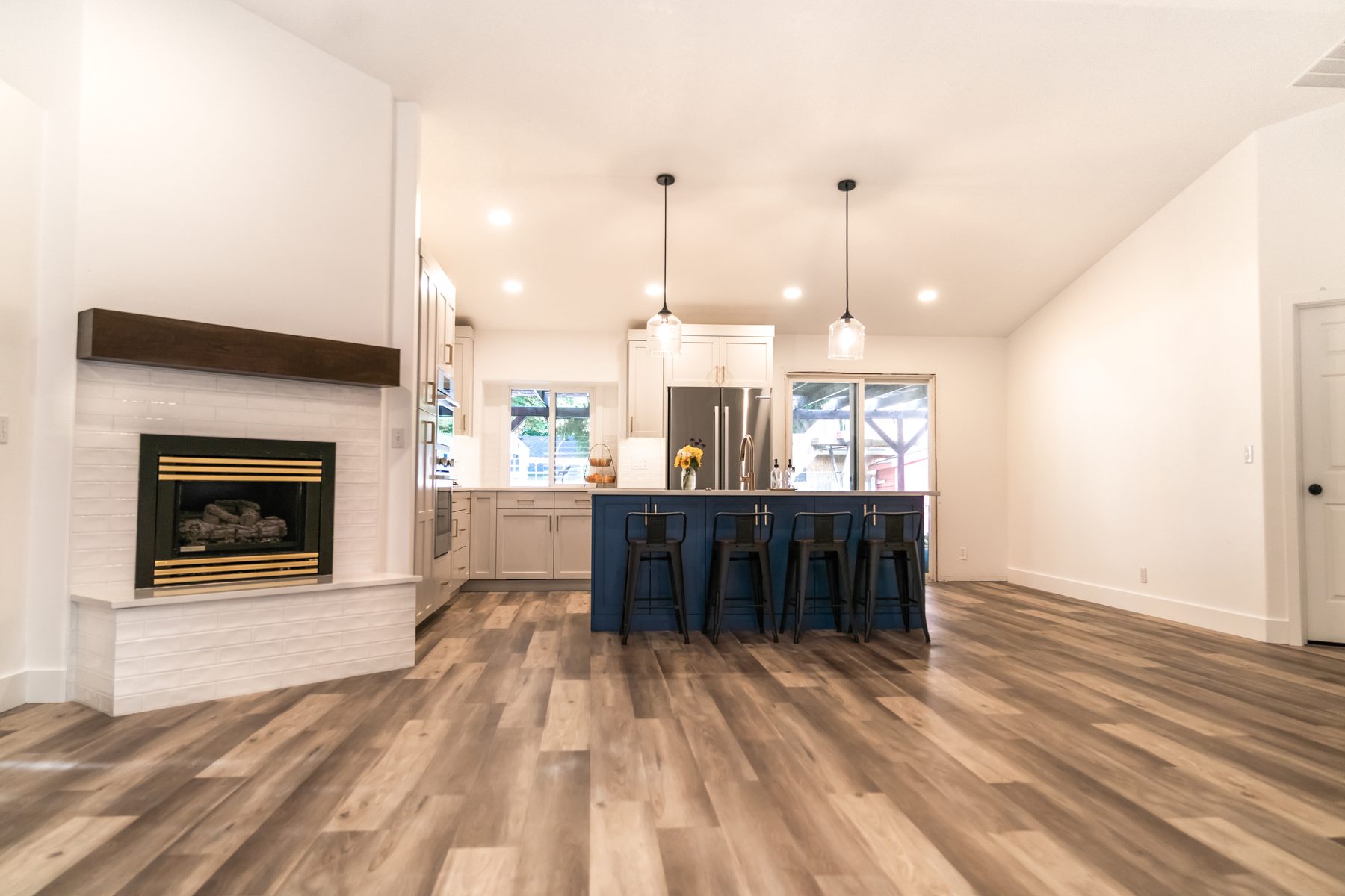 Open kitchen and living room remodel with white shaker cabinets, a navy island with black stools, glass pendants, and a tiled fireplace with a walnut mantel