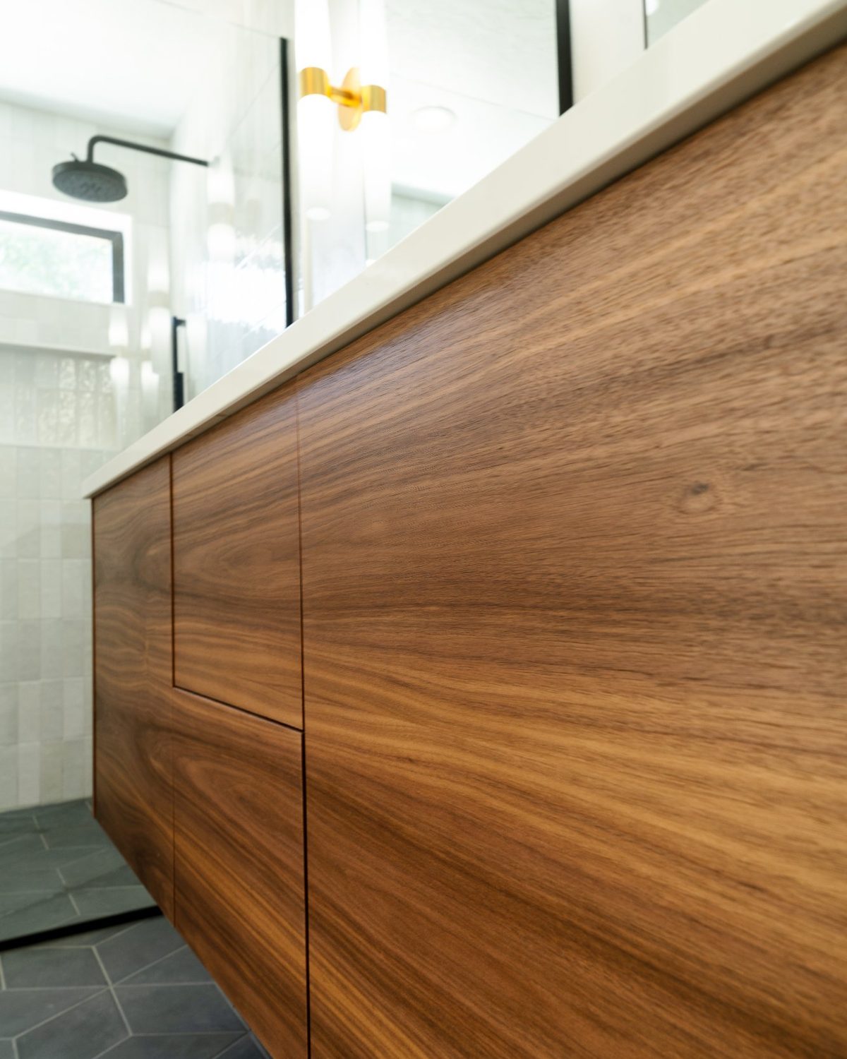 Grain-matched walnut vanity doors with a white quartz top, black rain head shower, and brass sconce behind